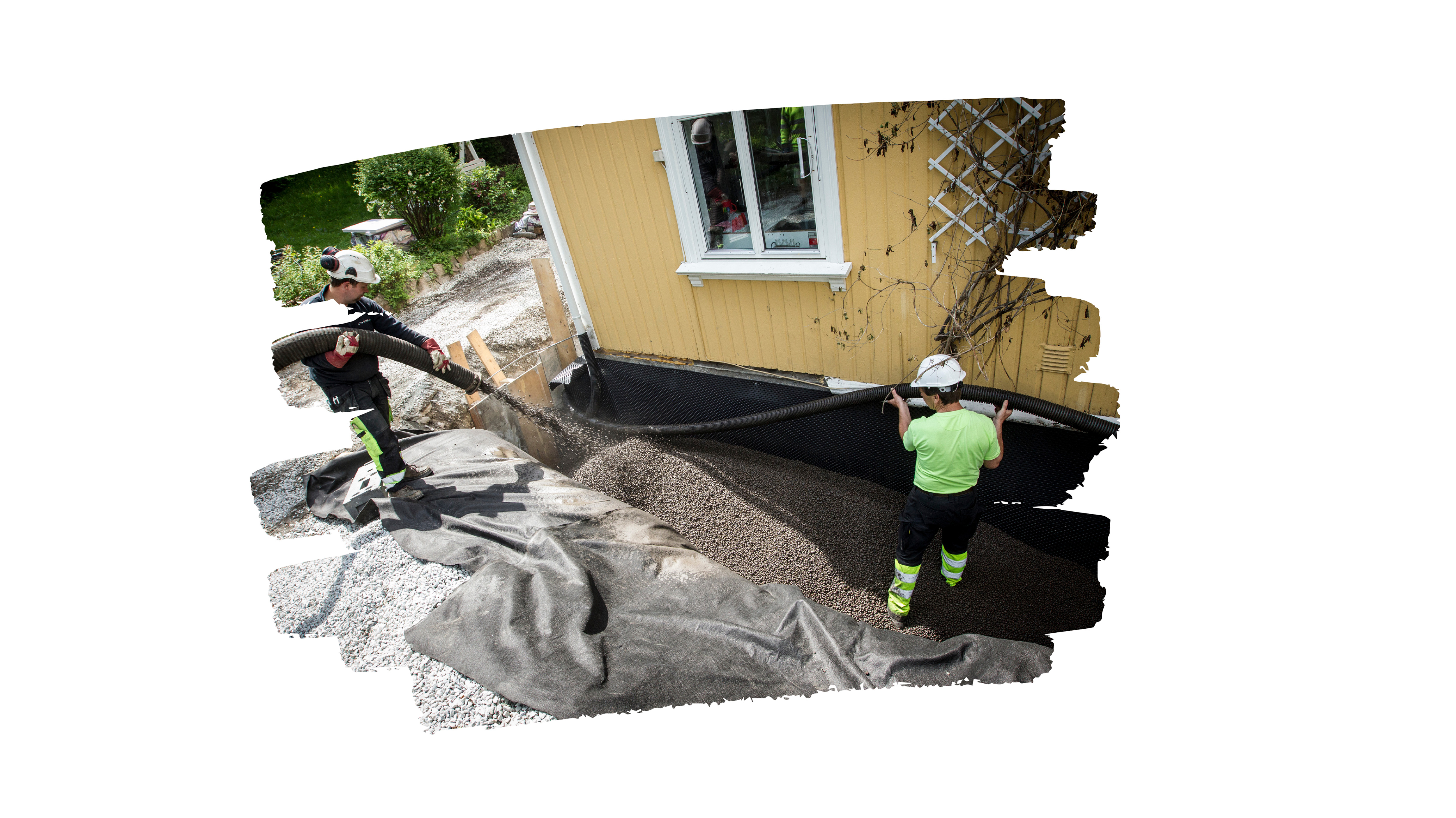 To arbeidere i verneutstyr blåser inn Leca-kuler rundt grunnmuren på en gul trehusbolig. Leca-kulene fyller et område dekket med fiberduk og dreneringsplate for å isolere og sikre god drenering. Området rundt viser grus, verktøy og en hage med planter og sykler i bakgrunnen.