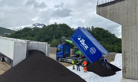 Blå Leca-lastebil tipper ut mørk lettklinker ved fundamentet til Ramfjordbrua. Tre personer i verneklær står ved fyllingen. Snødekt fjell sees i bakgrunnen under overskyet himmel.