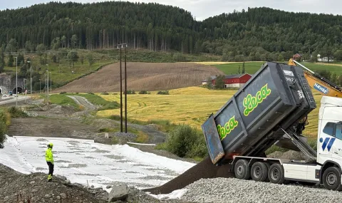En lastebil fra Leca tipper ut lettklinker på en interimvei ved Leistadkrysset på nye E6 Ranheim–Værnes. En anleggsarbeider i gul refleksdrakt står og overvåker arbeidet, mens en Hæhre-gravemaskin og jordbrukslandskap med åkre og skog sees i bakgrunnen.