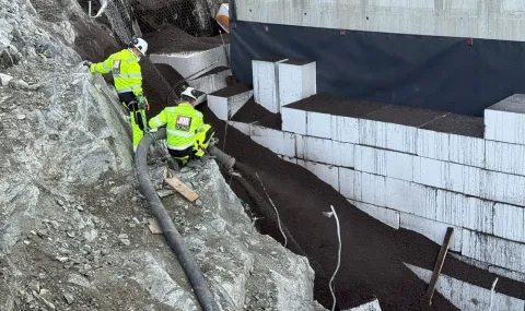 To anleggsarbeidere fra Jobzone blåser inn Leca® Lettklinker bak en betongkonstruksjon ved hjelp av fleksibelt slangeutstyr. Lettklinkeren fyller rommet bak EPS-blokker for å redusere jordtrykket mot byggets bakvegg i et bratt og utfordrende terreng.