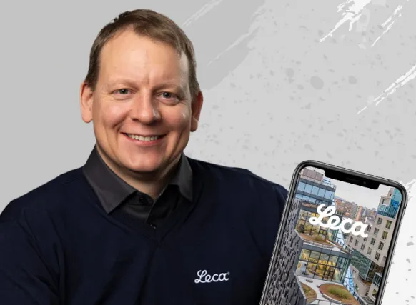 Smilende mann i mørk Leca-genser foran lys bakgrunn med grå penselstrøk. Ved siden av ham vises en mobiltelefon med et bilde av et moderne bygg og Leca-logoen på skjermen. Bildet illustrerer Leca og digitale verktøy for overvannshåndtering eller byggeprosjekter.