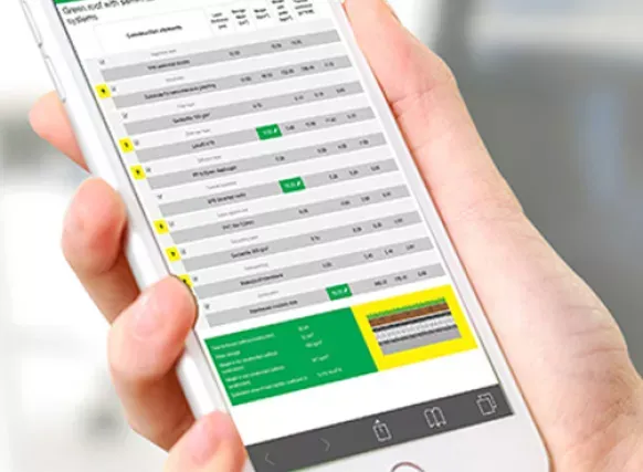 En hånd som holder en smarttelefon med skjerm som viser en teknisk tabell for grønne tak. Tabellen inneholder tekniske data og målinger for ulike lag i et taksystem, inkludert en illustrasjon nederst. Bildet illustrerer bruk av digitale verktøy for å planlegge eller dokumentere byggetekniske løsninger.