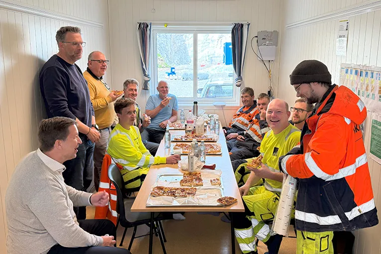 Leca- og Skanska-ansatte nyter pizza sammen i en brakkerigg ved Hålogalandsveien, med godt humør og samtaler rundt bordet.