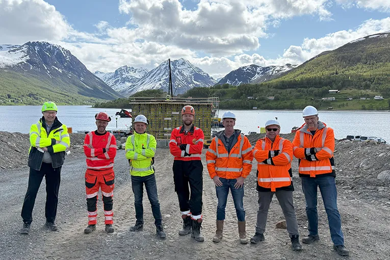 Gruppe i verneklær og hjelmer poserer foran brokar ved Ramfjorden i Tromsø, med fjell og vann i bakgrunnen