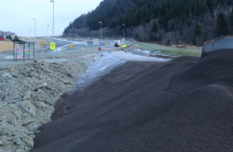 Stor haug med mørk Leca Lettklinker på anleggsområde ved Leistadkrysset, med veiskilt og trafikk i bakgrunnen.