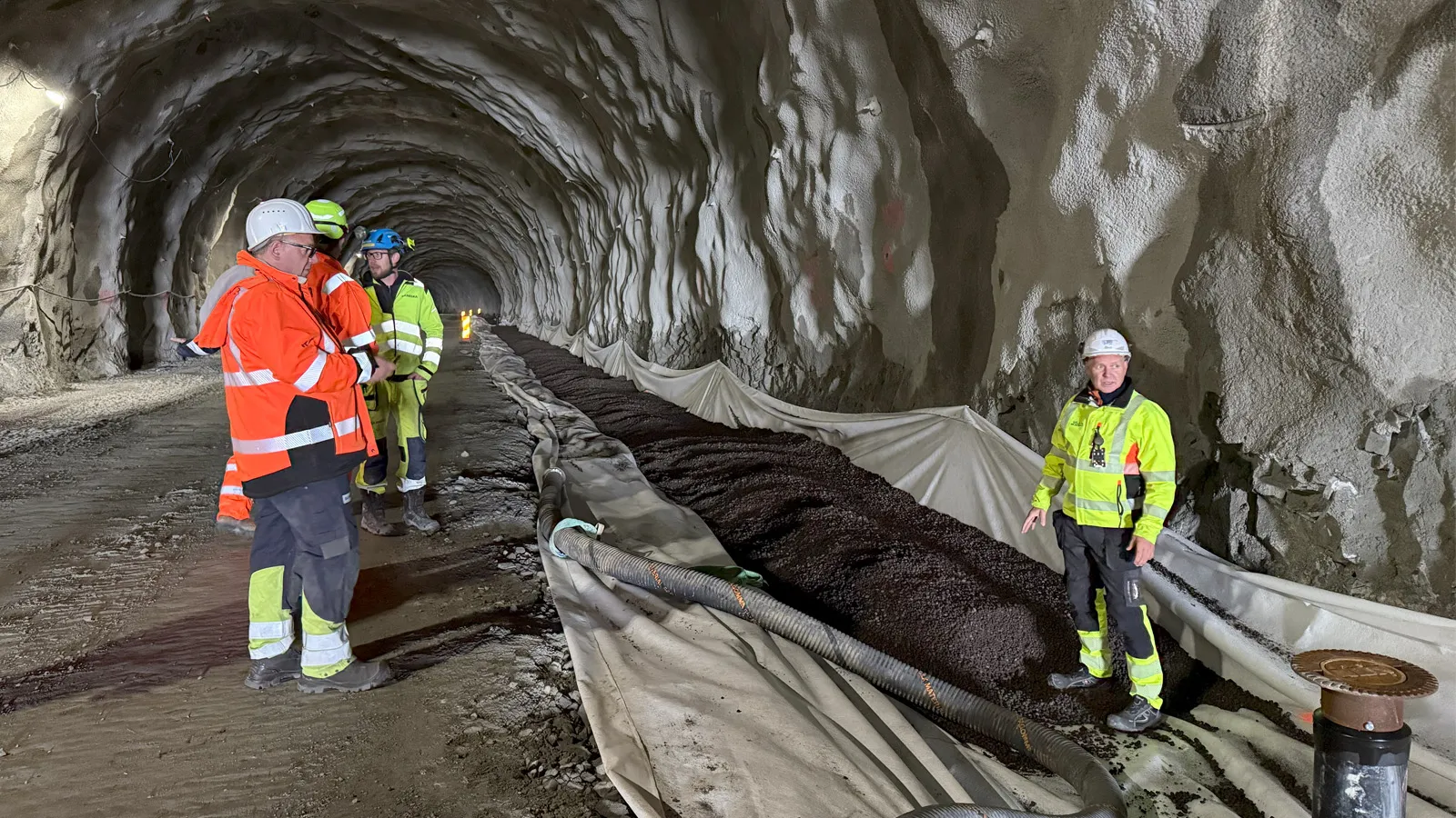 Arbeidere fra SKANSKA og Leca inspiserer utlegging av Leca Lettklinker som isolasjonslag rundt vannledninger inne i Brattfjelltunnelen, en del av Hålogalandsvegen-prosjektet. Lettklinkeren ligger på fiberduk og viser et bærekraftig og effektivt tiltak i tunnelkonstruksjon.