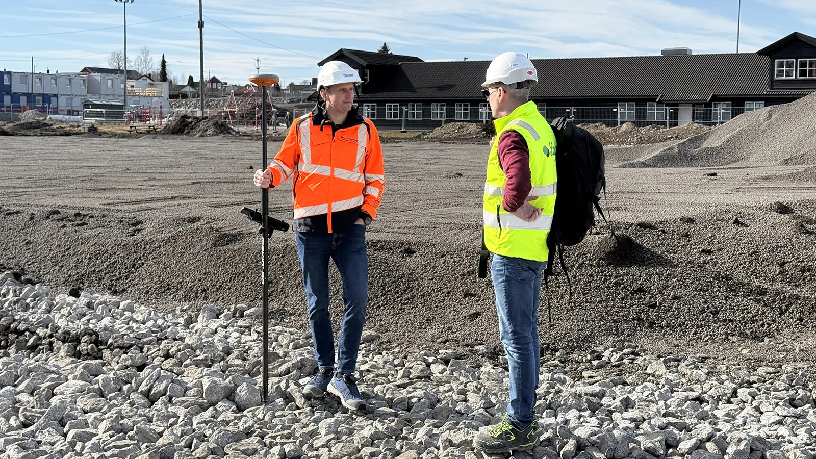 To anleggsarbeidere med hjelm og refleksvester i samtale på byggeplassen for den nye kunstgressbanen i Stange. I forgrunnen ligger Leca® Lettklinker og pukk som grunnlag for banen