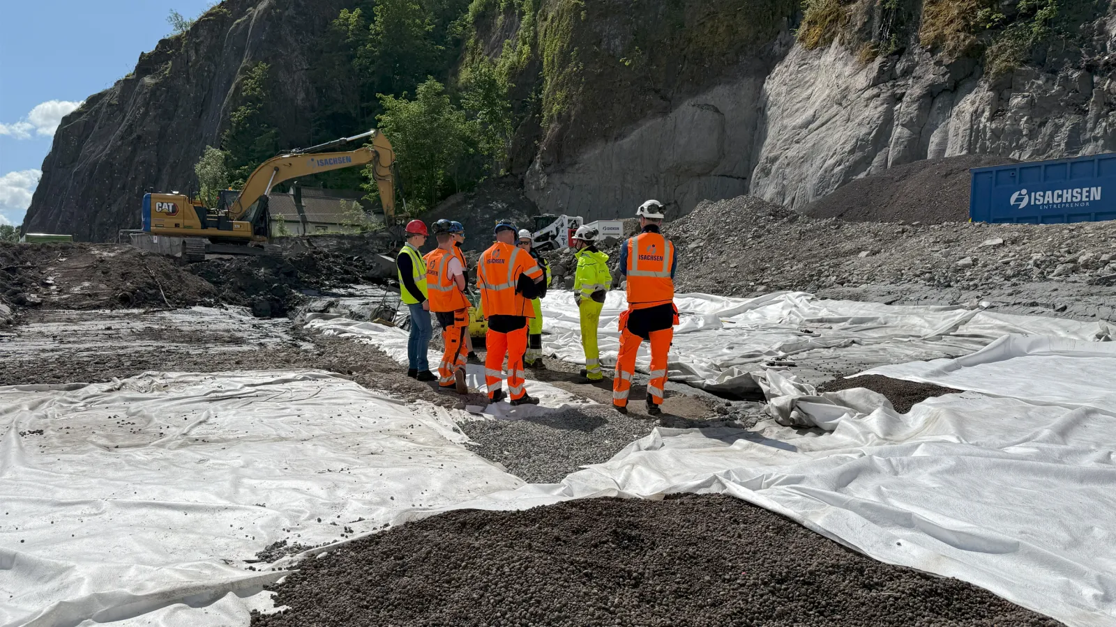Anleggsarbeidere fra Isachsen og samarbeidspartnere inspiserer utlegging av Leca® Lettklinker på byggeplassen i Holmestrand sentrum. Bildet viser geotekstil og lettklinker på bakken, med fjellskjæring og gravemaskin i bakgrunnen.