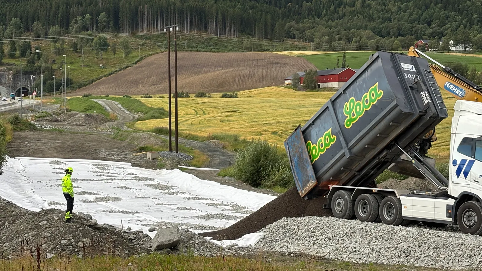 En lastebil fra Leca tipper ut lettklinker på en interimvei ved Leistadkrysset på nye E6 Ranheim–Værnes. En anleggsarbeider i gul refleksdrakt står og overvåker arbeidet, mens en Hæhre-gravemaskin og jordbrukslandskap med åkre og skog sees i bakgrunnen.