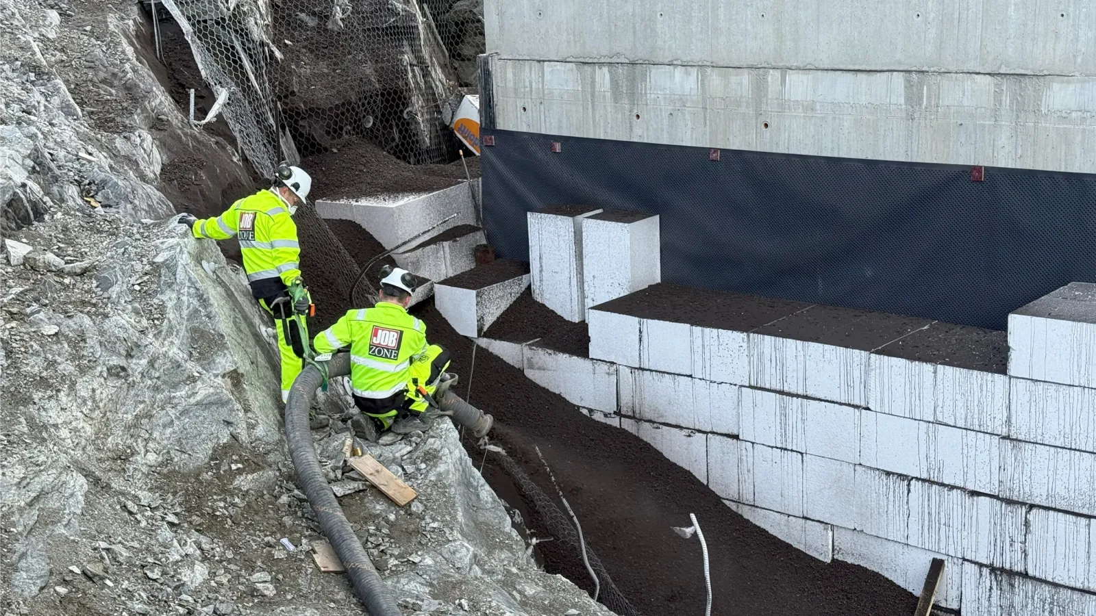 To anleggsarbeidere fra Jobzone blåser inn Leca® Lettklinker bak en betongkonstruksjon ved hjelp av fleksibelt slangeutstyr. Lettklinkeren fyller rommet bak EPS-blokker for å redusere jordtrykket mot byggets bakvegg i et bratt og utfordrende terreng.