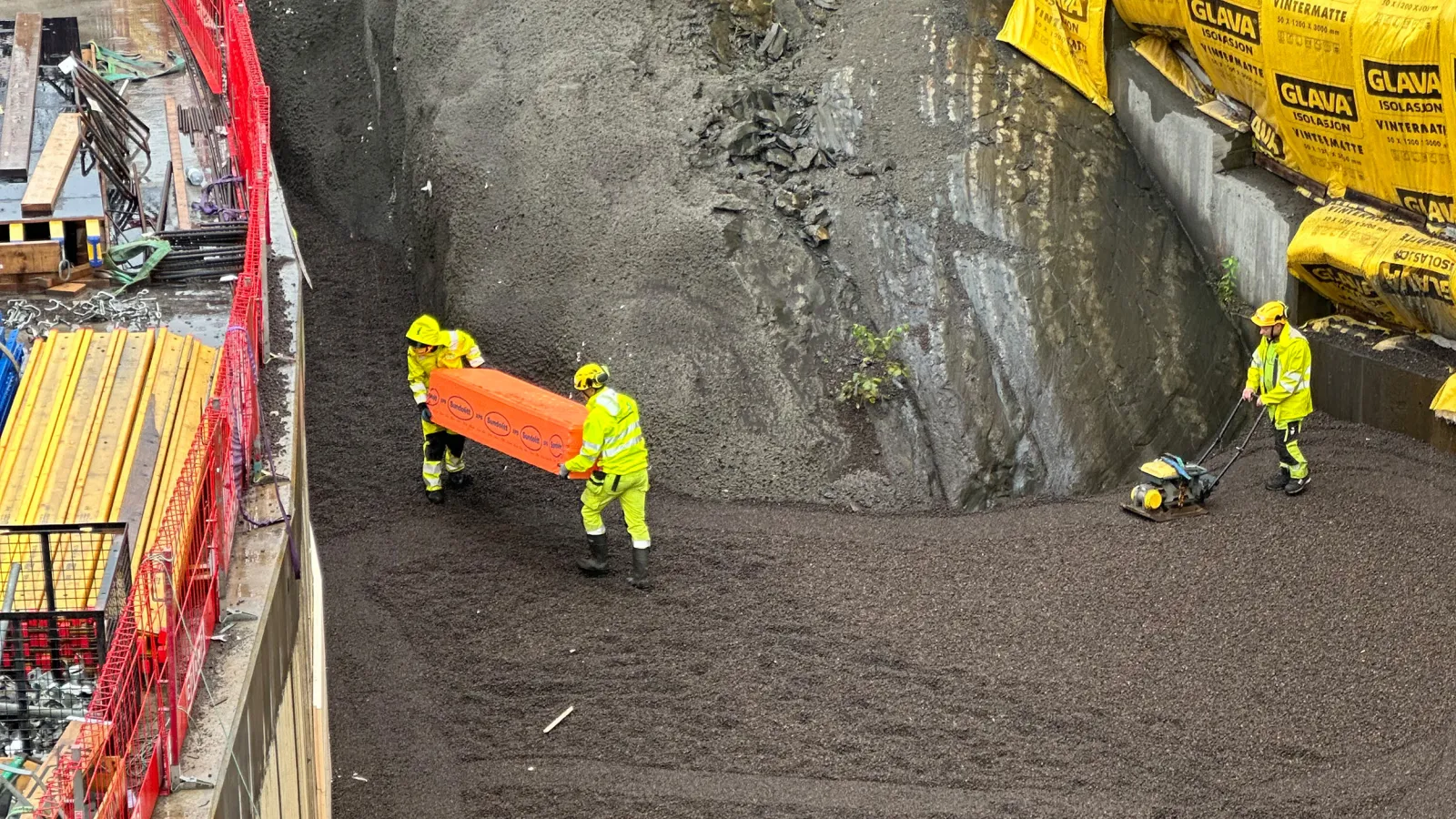 Arbeidere i gule verneklær legger ut og komprimerer Leca® Lettklinker i en 11 meter dyp grøft på Fornebu stasjon.