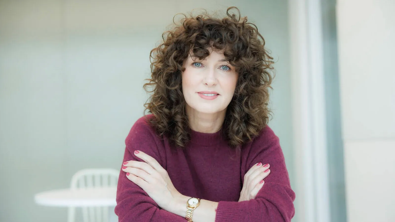 Portrett av Ilona Miszczak, ny administrerende direktør i Leca. Hun sitter ved et hvitt bord med armene i kors og ser rett mot kameraet med et vennlig uttrykk. Hun har mørkt, krøllete hår og har på seg en vinrød genser. Bakgrunnen er lys og nøytral, med en hvit stol synlig i bakgrunnen