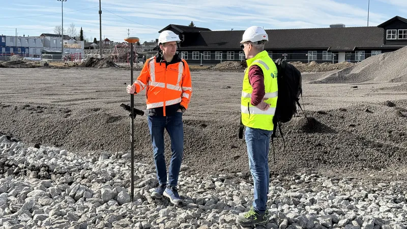 To anleggsarbeidere med hjelm og refleksvester i samtale på byggeplassen for den nye kunstgressbanen i Stange. I forgrunnen ligger Leca® Lettklinker og pukk som grunnlag for banen