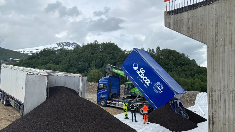 Blå Leca-lastebil tipper ut mørk lettklinker ved fundamentet til Ramfjordbrua. Tre personer i verneklær står ved fyllingen. Snødekt fjell sees i bakgrunnen under overskyet himmel.