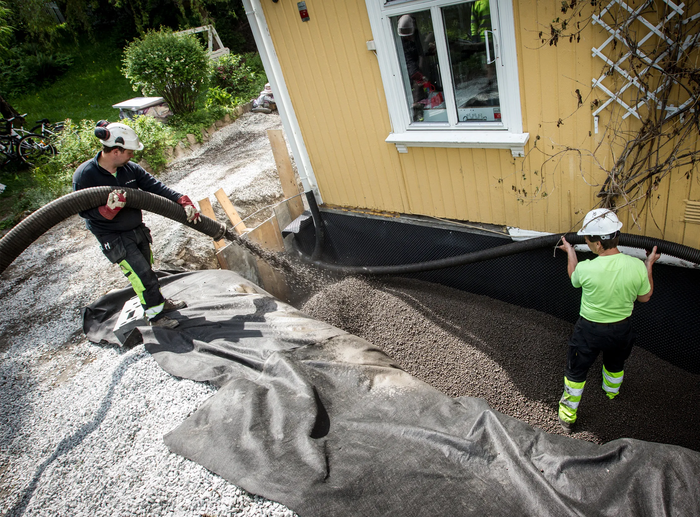 To arbeidere blåser inn Leca lettklinker som drenerende masse langs grunnmur på en enebolig.