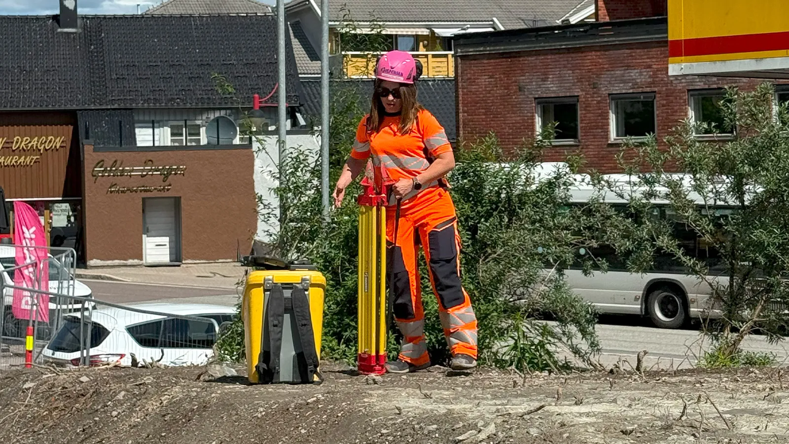 vinne i oransje arbeidsklær og rosa hjelm håndterer oppmålingsutstyr ved byggeplass i Holmestrand sentrum, med trafikk og bygninger i bakgrunnen.