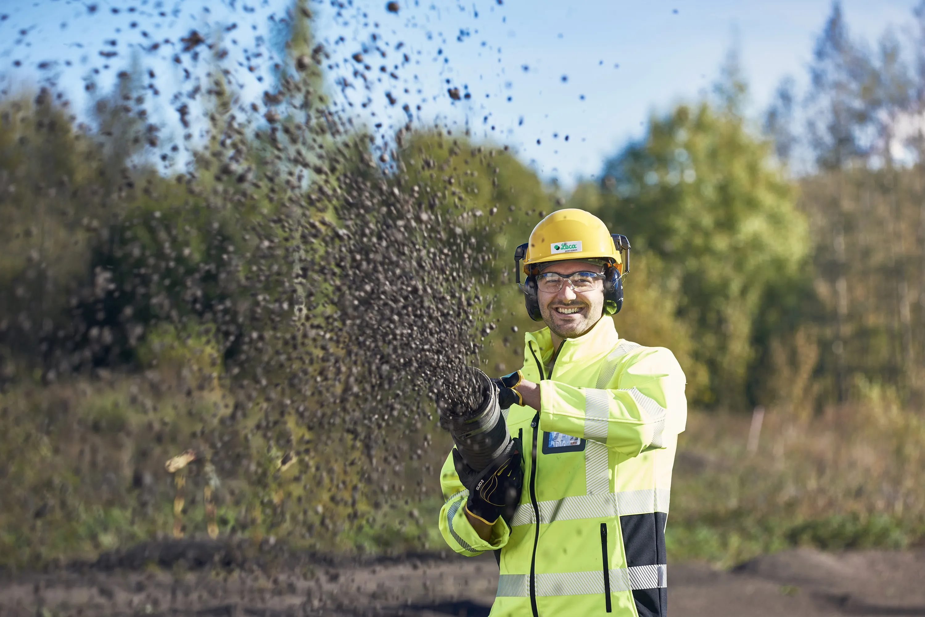En arbeidstaker med gul vernehjelm og gul refleksjakke blåser ut Leca-kuler fra en slange i høy hastighet. Han smiler mens kulene spruter utover i forgrunnen. I bakgrunnen er det trær og blå himmel på en utendørs byggeplass.