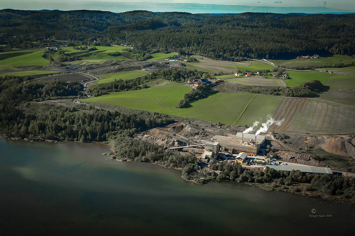 Luftfoto av Leca-fabrikken på Rælingen, omgitt av grønne jorder og skog, med produksjonsanlegg ved vannkanten og damp som stiger fra fabrikkpipene