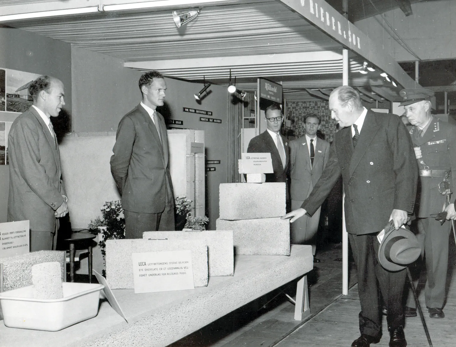 Sort-hvitt-bilde fra 1955 der kong Olav V besøker Leca sin stand på Bygg Reis Deg, og undersøker lettklinkerblokker sammen med representanter fra selskapet.