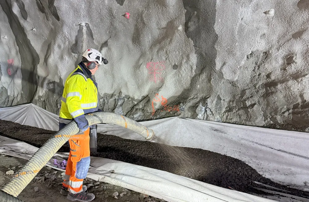 Arbeider i verneutstyr blåser inn Leca Lettklinker som isolasjonslag i grøft inne i Gullesfjordtunnelen. Materialet blåses støvfritt over fiberduk for frostsikring av rør.