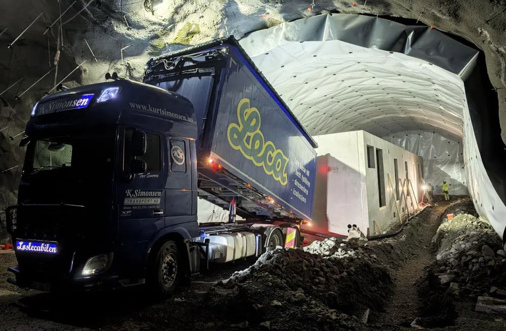 Leca-blåsebil fra K. Simonsen tipper lettklinker inne i tunnel med prefabrikkert teknisk bygg. Arbeidet skjer under belysning i kveldsmørke.