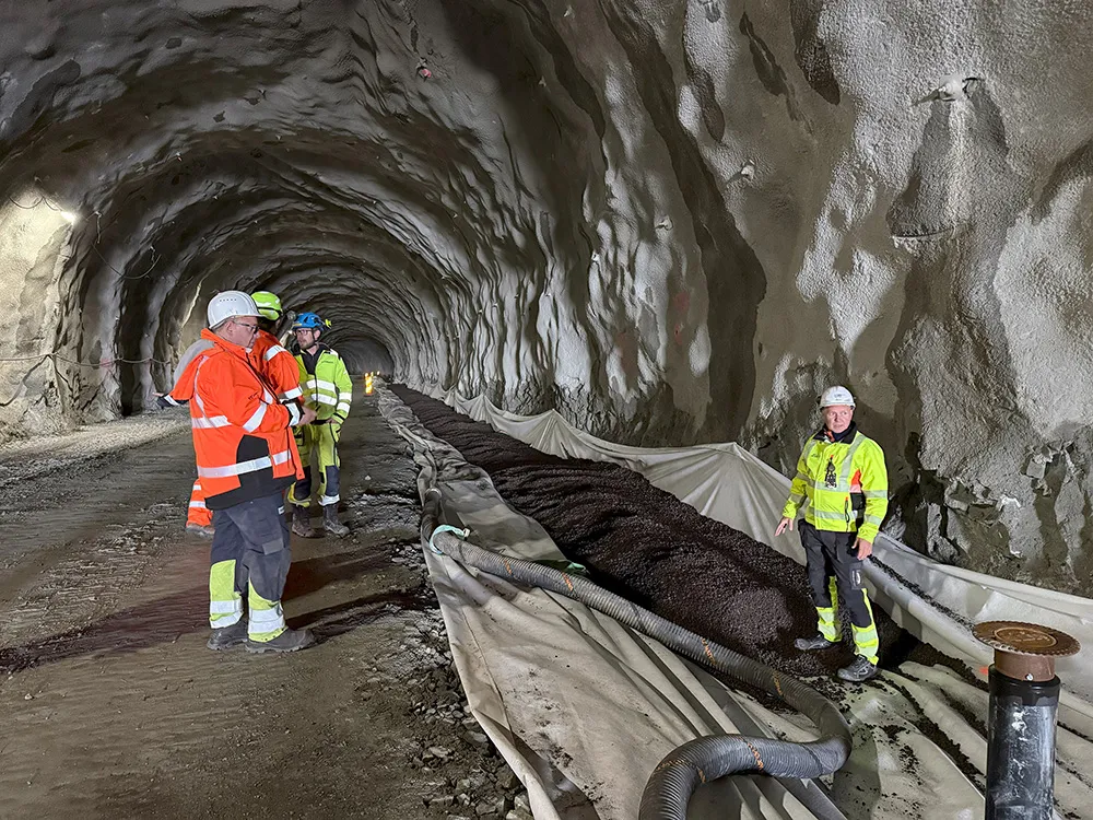 Flere personer i verneklær og hjelm inspiserer tunnelarbeid der Leca Lettklinker er blåst inn i grøft som frostisolasjon