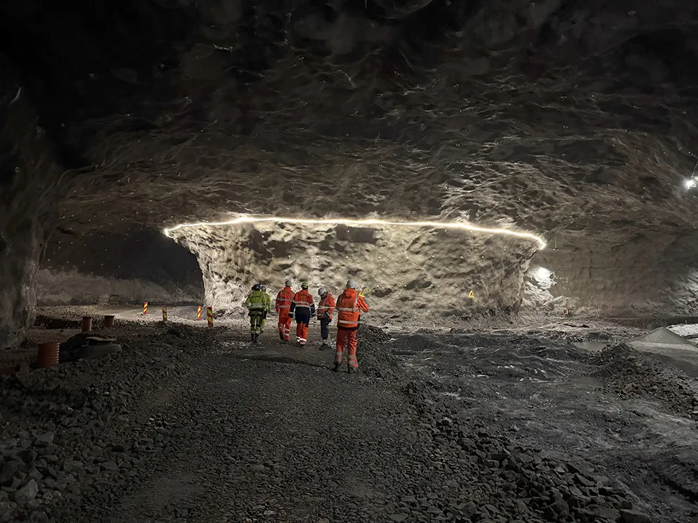 Gruppe i oransje og gule verneklær går innover i en stor tunnel med ujevne fjellvegger og arbeidslys.