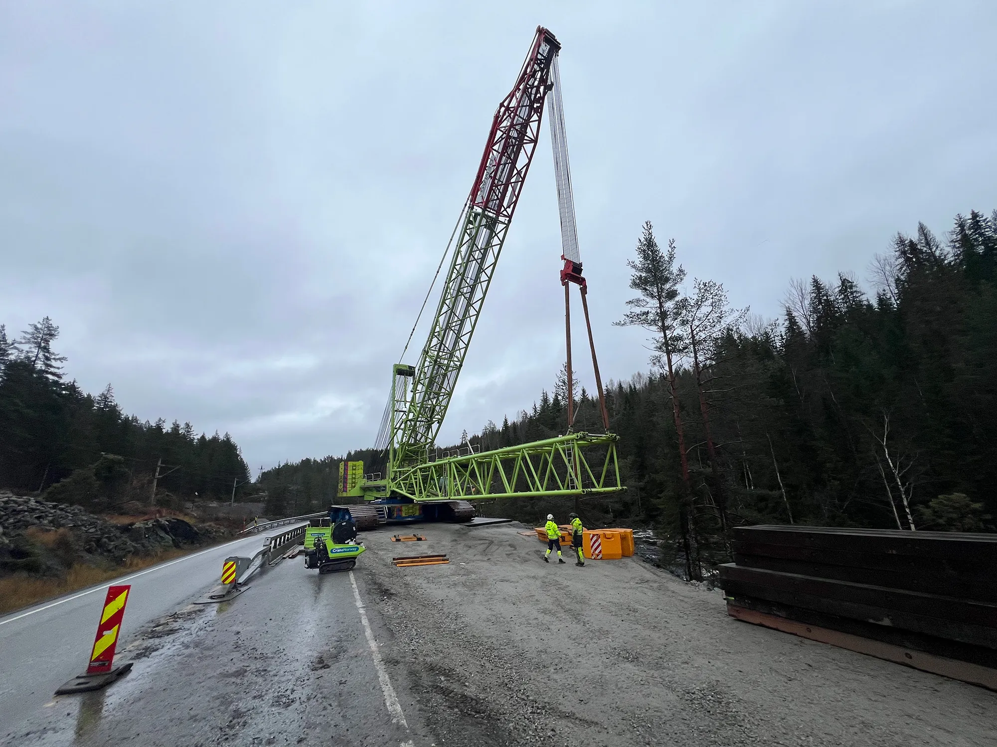 Stor grønn og rød beltekran rigget opp langs veien i skogsområde. To arbeidere i refleksklær står i nærheten mens kranen forbereder tungløft.
