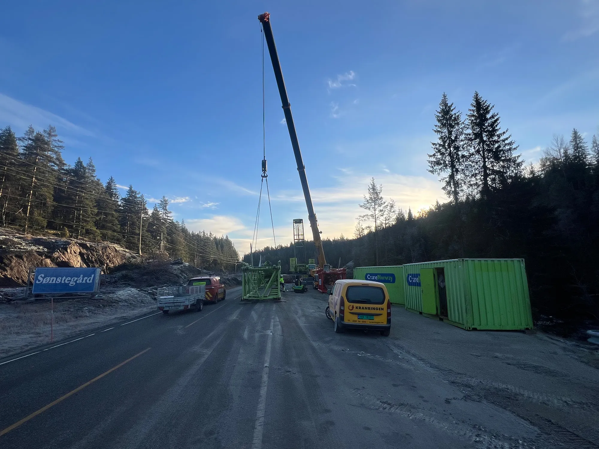 Mobilkran løfter en grønn stålkonstruksjon på en midlertidig riggplass langs veien. Servicebiler og containere står parkert, med skog og lav sol i bakgrunnen.