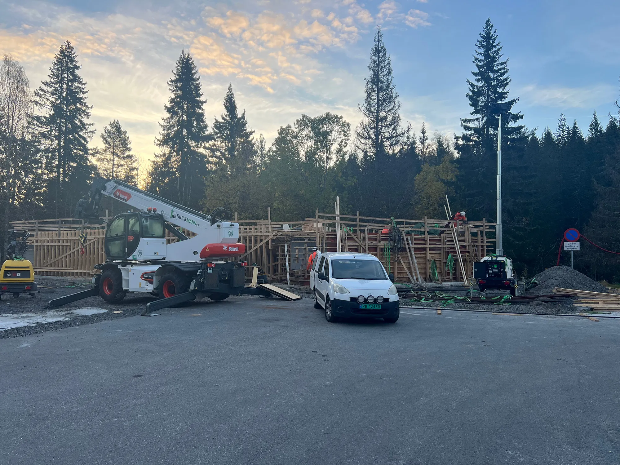 Anleggsområde med forskaling i tre, gravemaskin og servicebil foran en konstruksjon under oppføring. Arbeidere i verneutstyr er synlige, med skog og kveldshimmel i bakgrunnen.