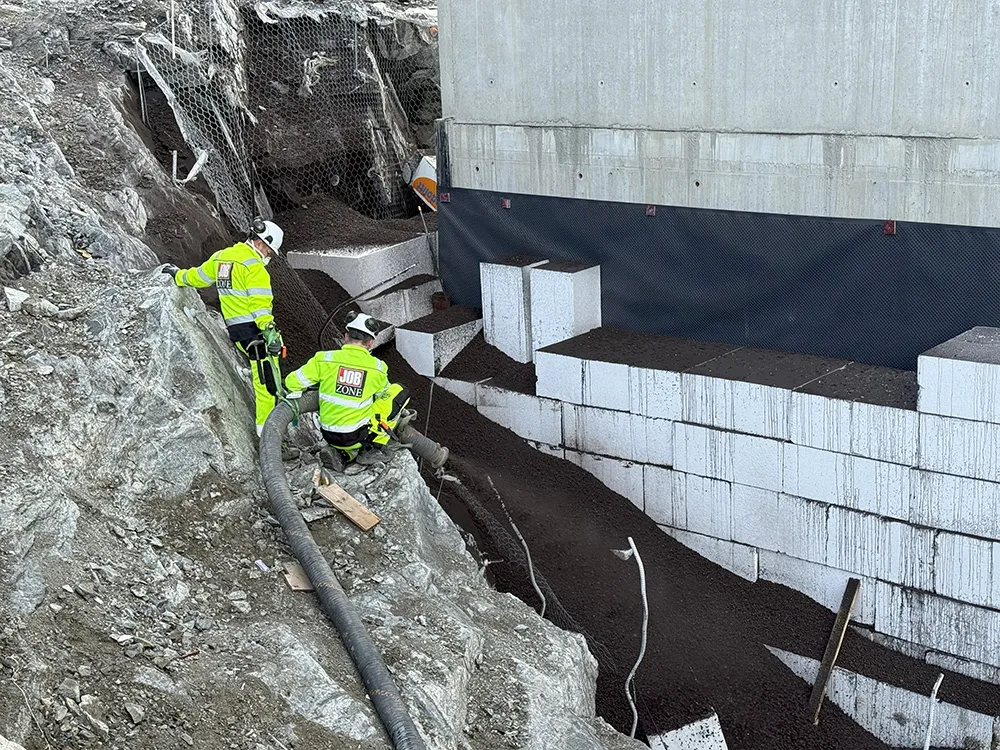 To arbeidere blåser Leca Lettklinker inn bak en støttemur av isoporelementer inntil betongvegg i skrånende terreng.