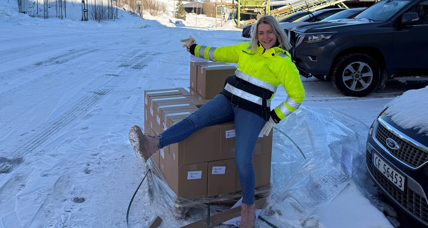 Smilende person i refleksjakke sitter på paller med esker med Leca Lettklinker teknisk håndbok, på et vinterlig lagerområde.