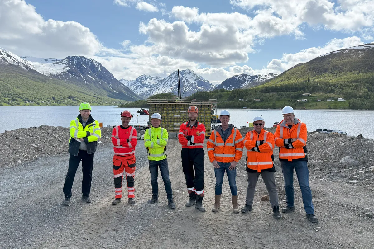 Gruppe med prosjektmedarbeidere i vernetøy og hjelmer foran Ramfjorden, med snødekte fjell i bakgrunnen og brofundament under bygging i midten av bildet.