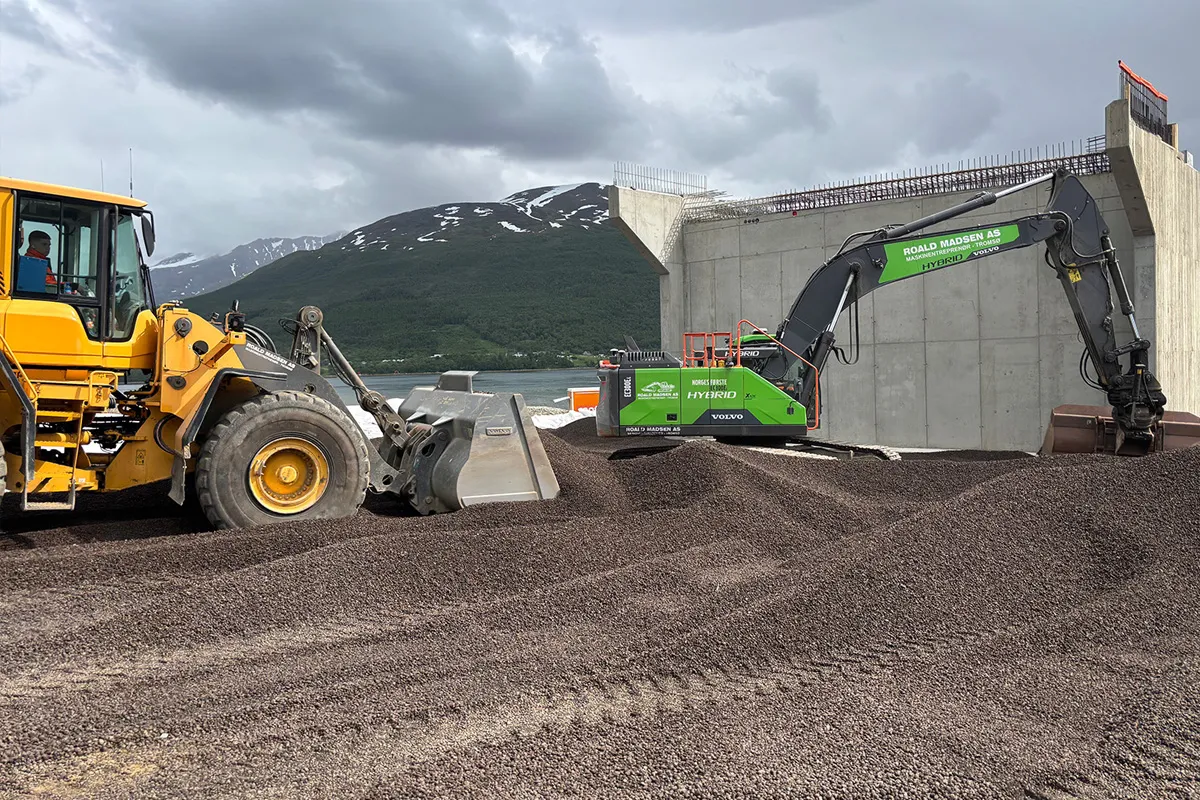 Gul hjullaster og grønn hybridgravemaskin som planerer Leca® Lettklinker foran brofundamentet ved Ramfjorden, med fjell og vann i bakgrunnen.