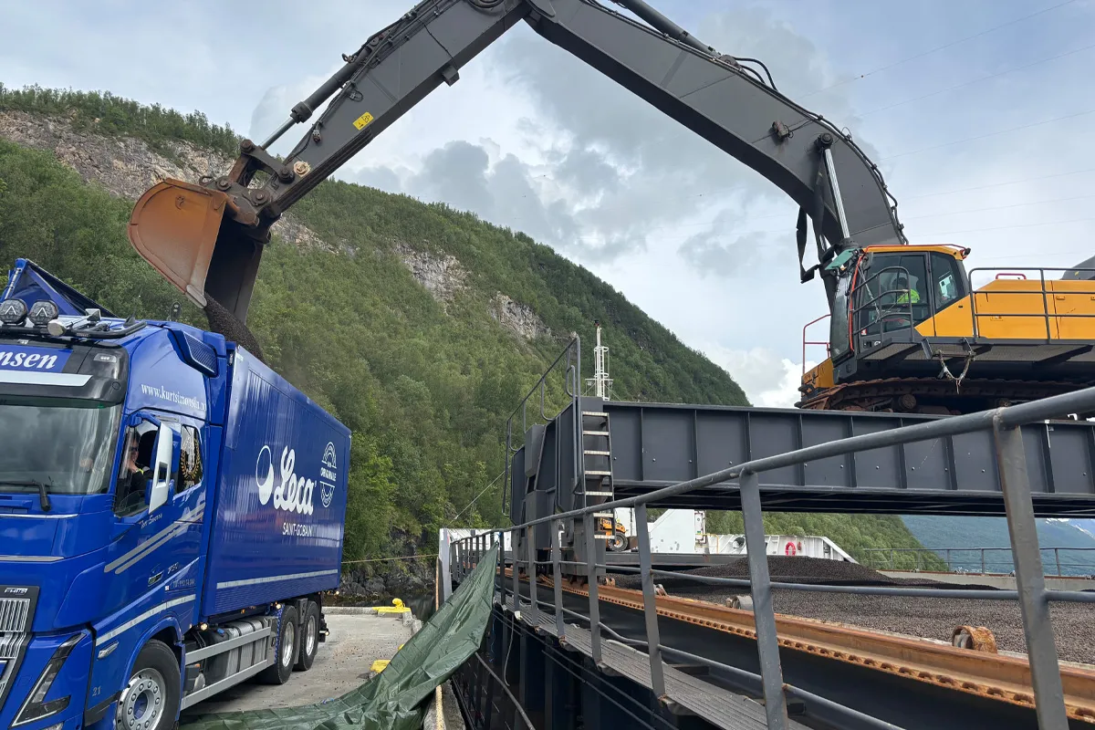 Gravemaskin som laster Leca® Lettklinker fra båt over i blå Leca-lastebil ved kai, med skogkledd fjellside i bakgrunnen.