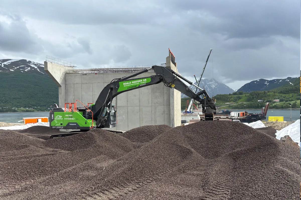 Grønn hybridgravemaskin fra Roald Madsen AS planerer Leca® Lettklinker foran brofundamentet ved Ramfjorden, med fjell og vann i bakgrunnen under overskyet himmel.
