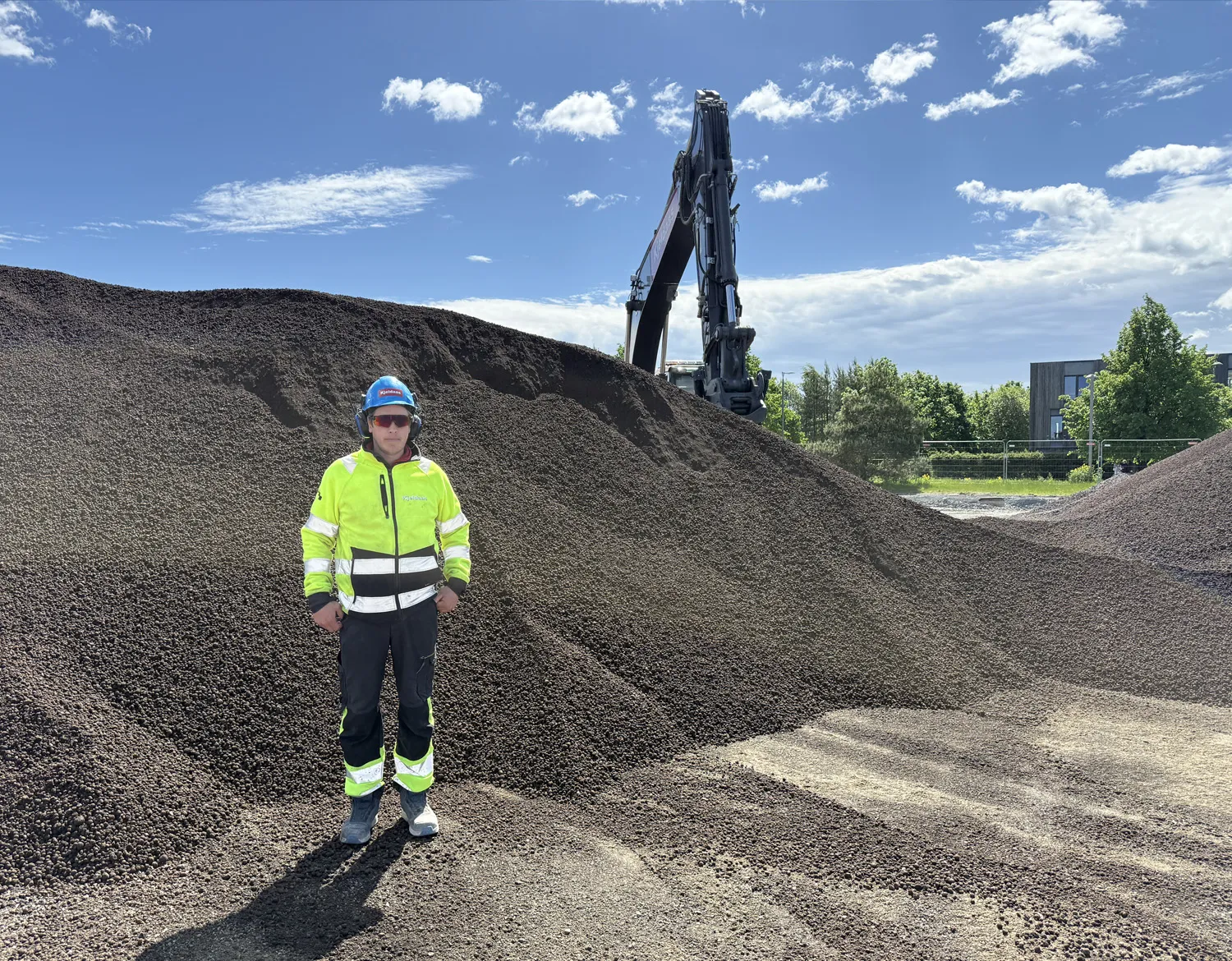 Person i verneutstyr står foran store hauger med gjenbrukt Leca® Lettklinker på anleggsområde, med gravemaskin i bakgrunnen.