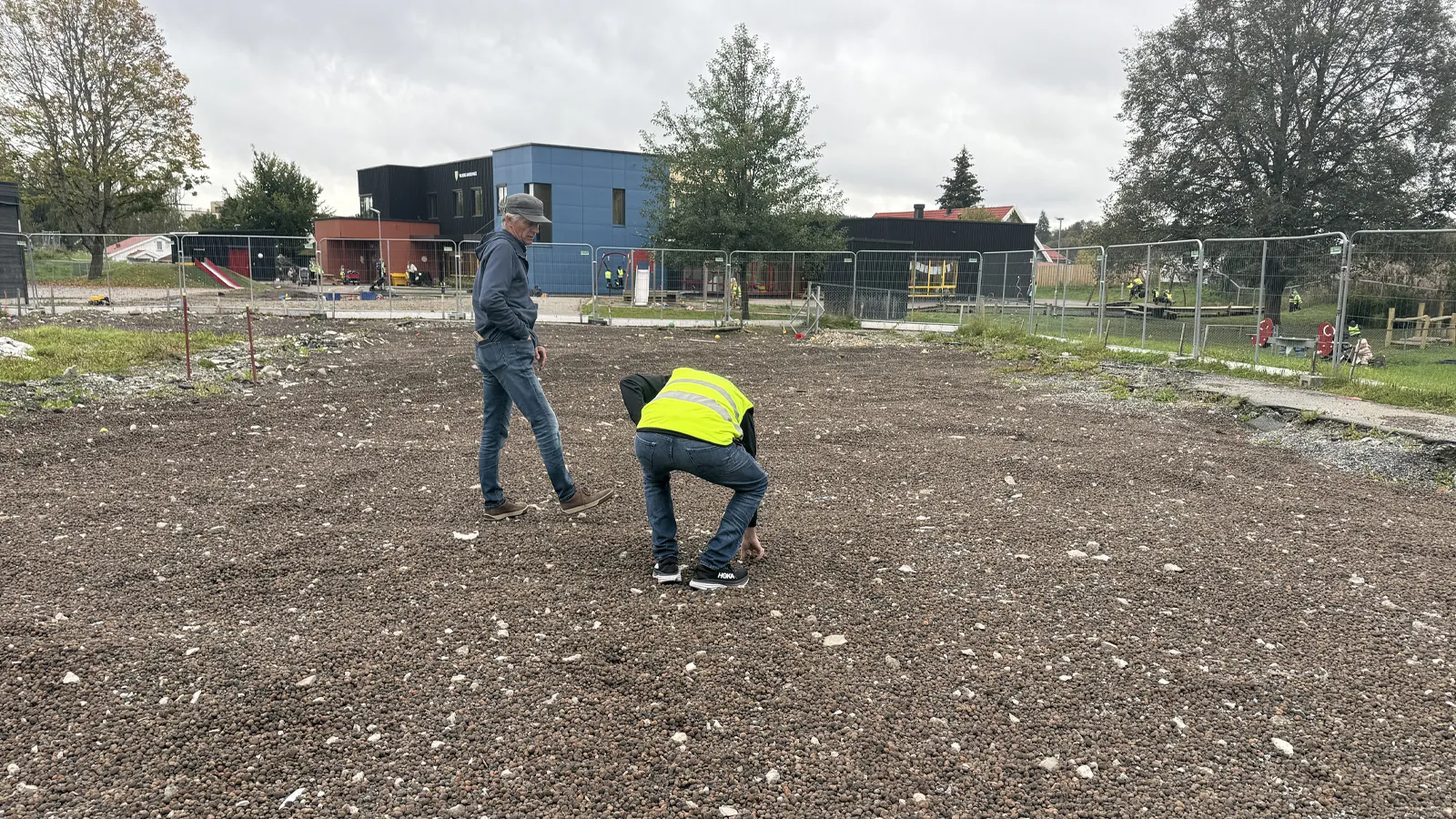 To personer står og snakker på byggeplassen ved Vilberg barnehage i Eidsvoll. Grunnen er dekket med gjenbrukt Leca, og barnehagebygg og lekeplass skimtes i bakgrunnen.