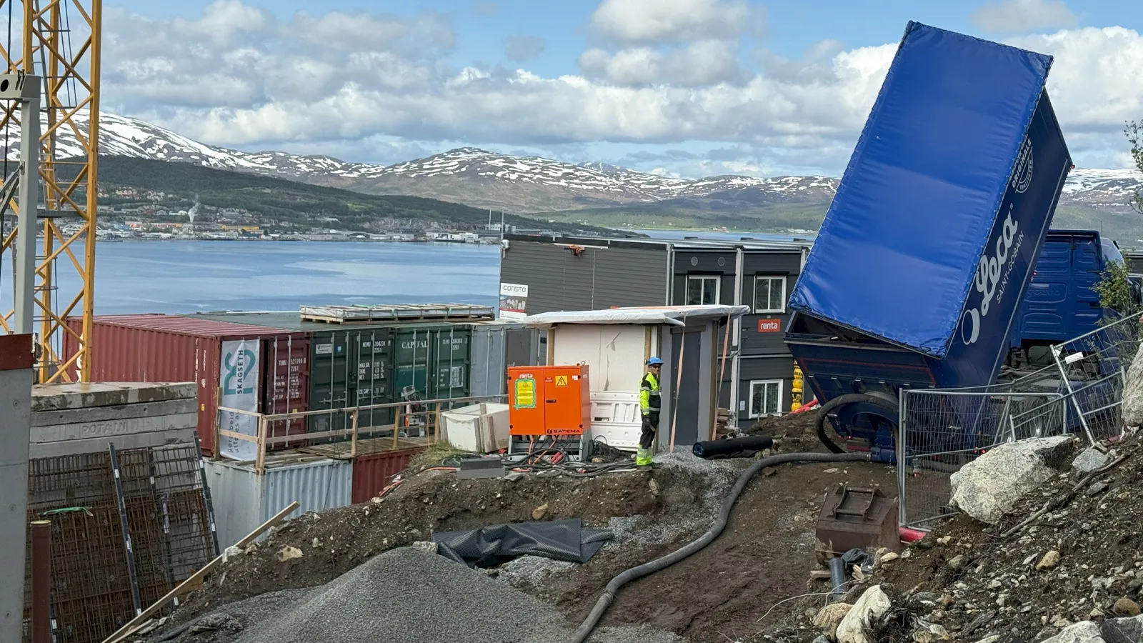 Blåsebil fra Leca tømmer lettklinker på byggeplass i Øvre Kroken med utsikt over fjorden og snødekte fjell. En arbeider følger prosessen tett i det kuperte terrenget.