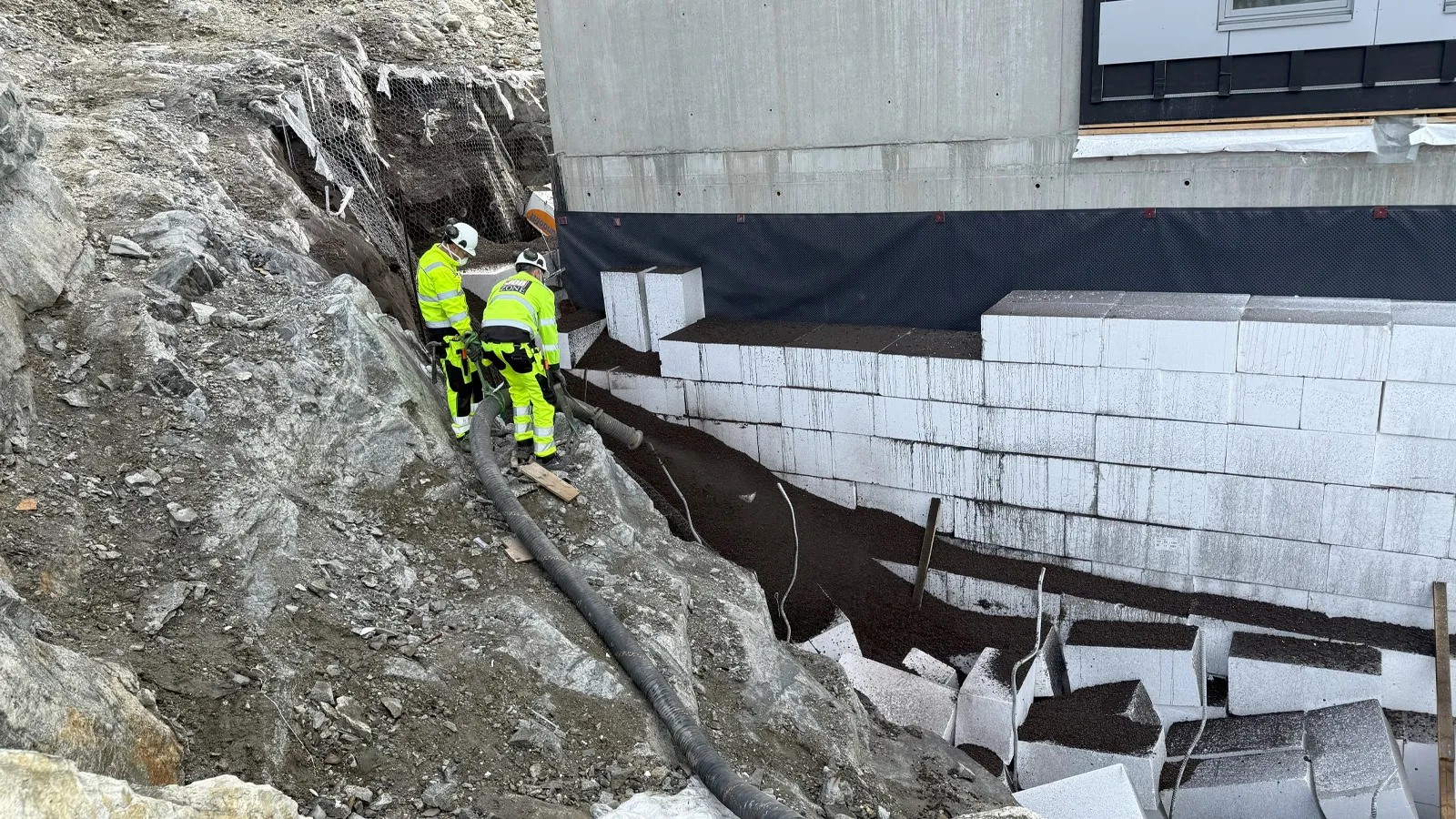 To arbeidere fra Jobzone blåser inn Leca® Lettklinker bak en betongvegg ved hjelp av blåseslange. Leca-massen fyller området bak EPS-blokker i en bratt fjellskråning.